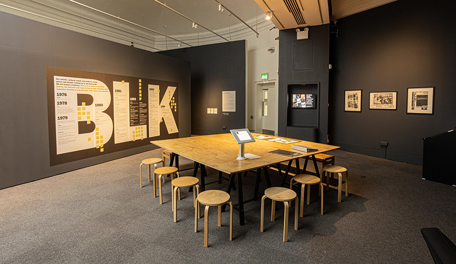 A photograph of a gallery space. The walls are painted dark grey and the floor is carpeted. On one wall there are three framed posters and a screen. The other wall has a timeline placed in the letters BLK in white font with yellow sticky notes. In the cen