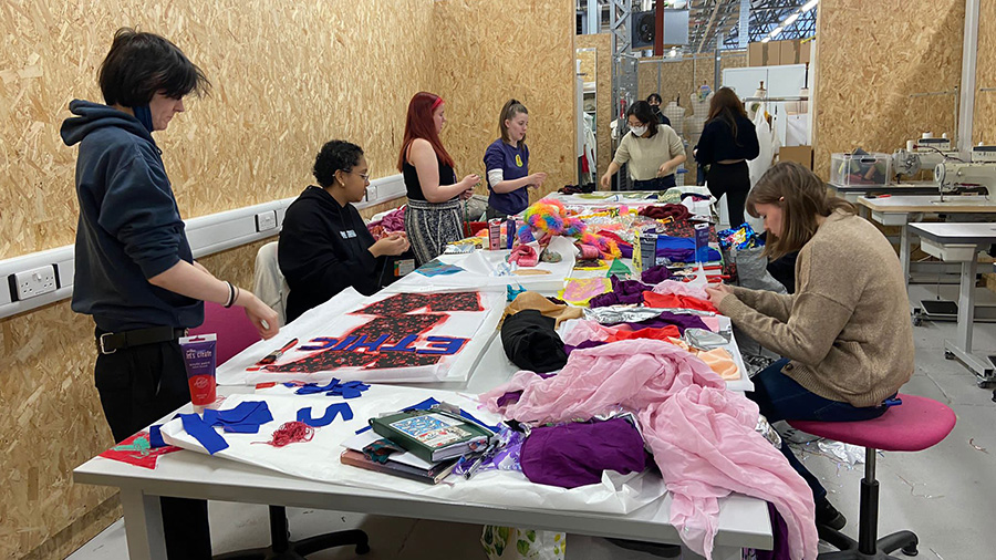 A group of people stand around a table creating artworks using fabric and paint.