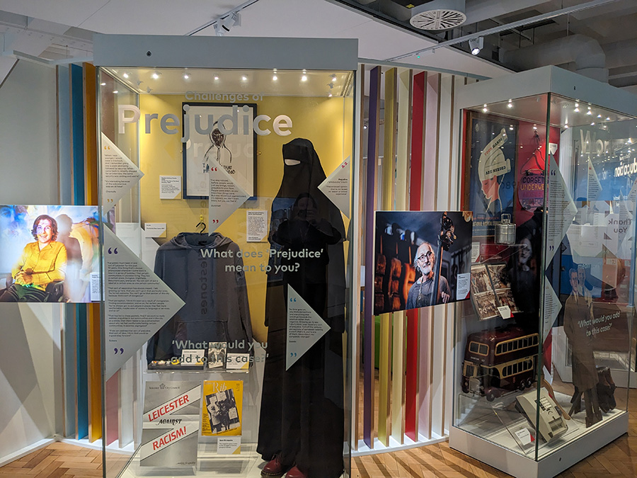 Interior of a museum. Glass display case labelled ‘prejudice’ contains several objects including a burqa and a hoodie