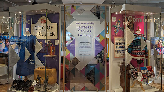 Interior of a museum. Glass display cases labelled ‘arrival’ and ‘community’ contain everyday items such as clothing and street signs