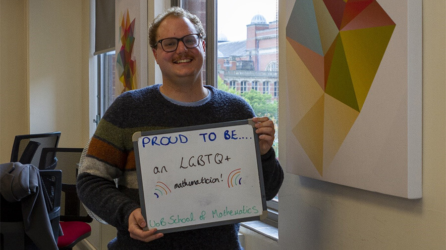 Rowland Seymour holding a sign saying Proud to be an LGBTQ+ mathematician, UoB School of Mathematics