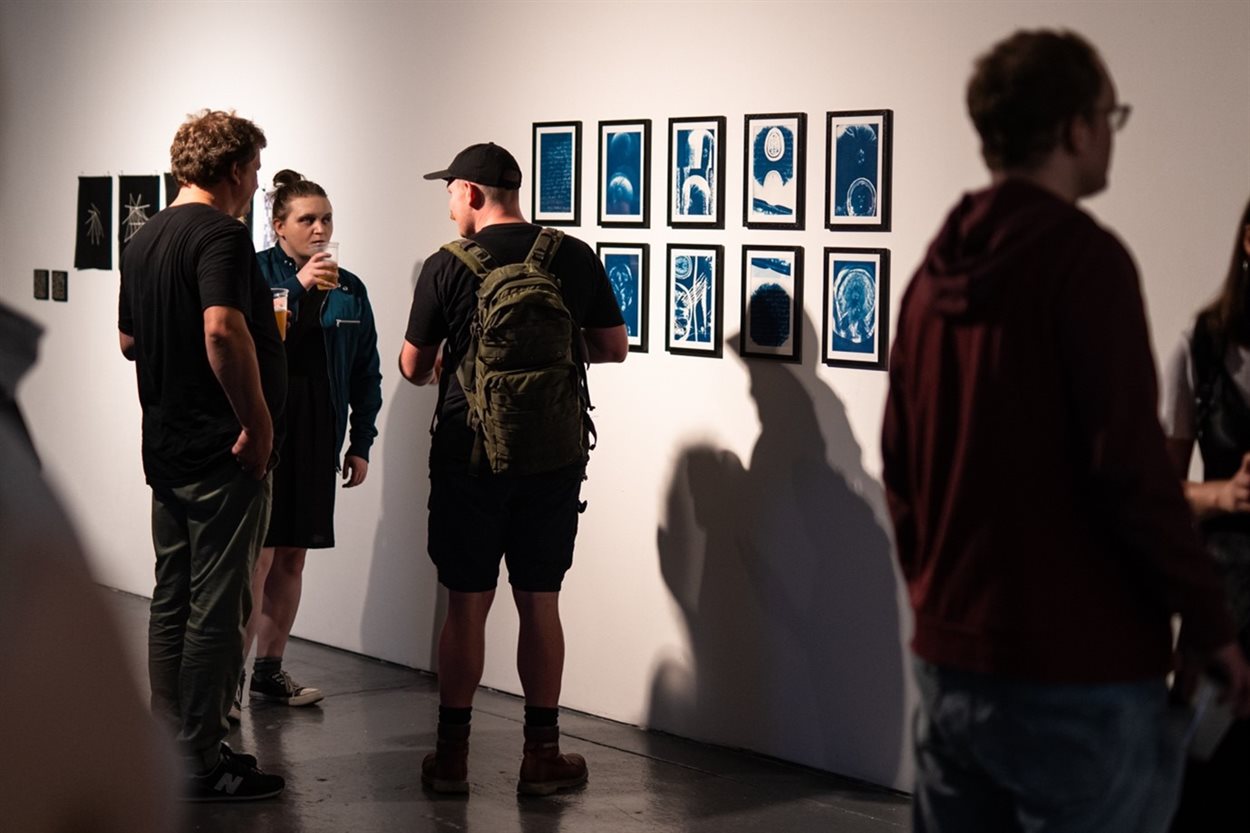 The photograph shows three young white men in their twenties or thirties wearing casual clothing such as shorts and drinking a beer whilst looking at a series of 10 blue and white cyanotype photography prints. The prints depict artwork by artist Mellissa Fisher about her condition papilledema. The artworks feature scans of her brain and photos of her eye alongside text not visible to the viewer.