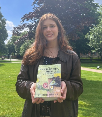 Student Olivia Eaton smiling in the sunshine on campus while holding a copy of Tamar Adler's 'The Everlasting Meal Cookbook'