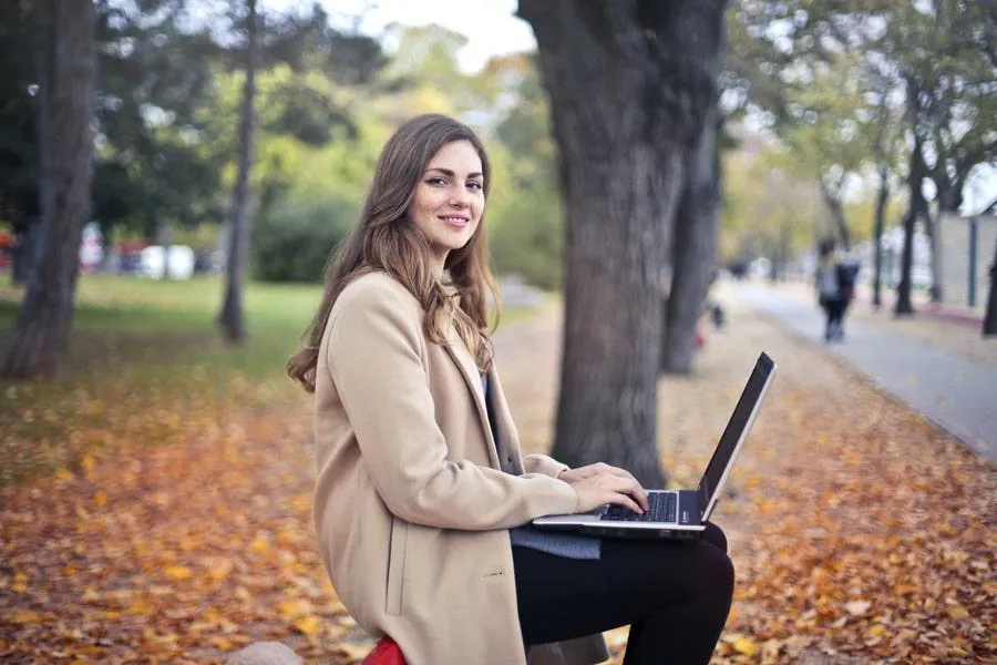 A young woman sitting on a park bench and smiling whilst using a laptop in an Autumn setting