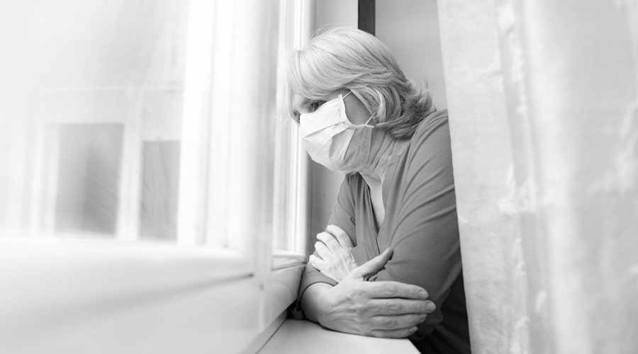 Sad senior woman looking out of her window with a face mask on