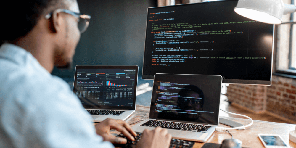 A person doing coding and looking at analytics across two laptops and one computer monitor at their desk.