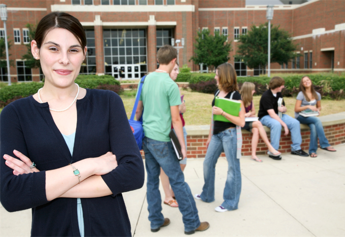 A teacher stands outside of a highschool, arms folded in front of their chest, while behind them stands two groups of students, one group of three stands and the second group of three speak between each other.