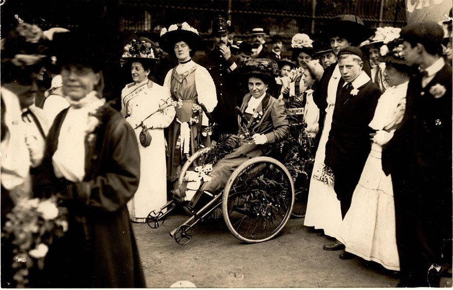 Billinghurst using "the machine" - her wheelchair at a Suffragette march. The wheelchair is decorated with flowers, and she wears a long dress and a big hat, and is surrounded by women similarly dressed.