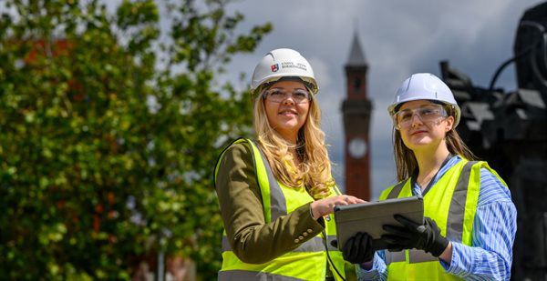 Two women in hi-vis jackets and hard hats stare ahead, with a large clock-tower in the background.