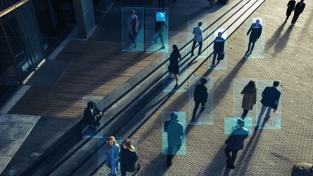 A crowd of people walking down a street are highlighted by blue boxes by an artificial intelligence surveillance system.