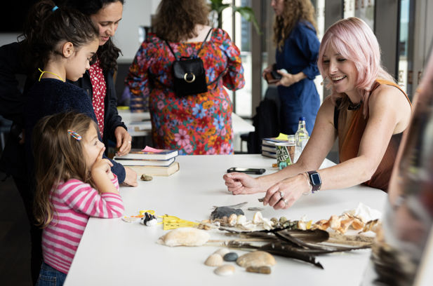 Professor. Alice Roberts wears a microphone as she shows a group of children some artifacts at a table.