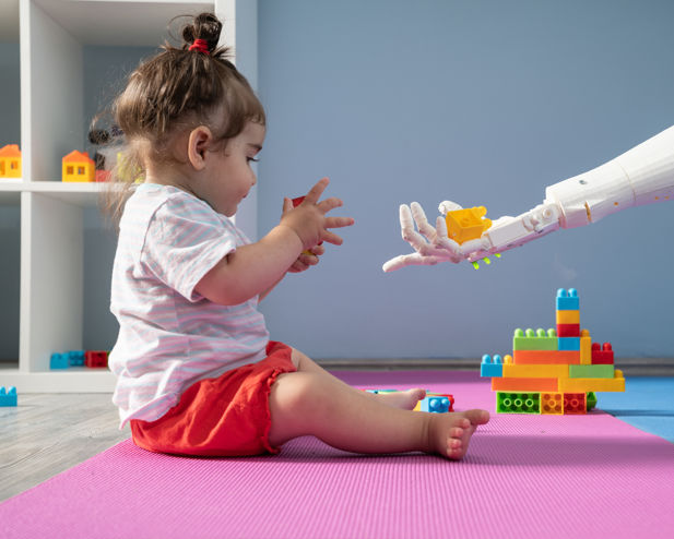 A white robot arm holds out plastic building blocks to a baby.