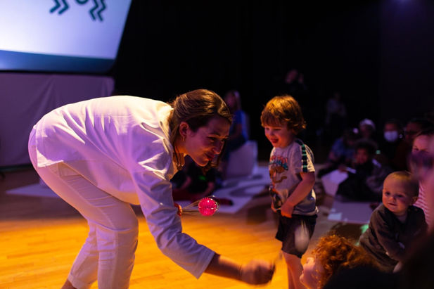 An adult woman reaches down with a toy to play with smiling young children.