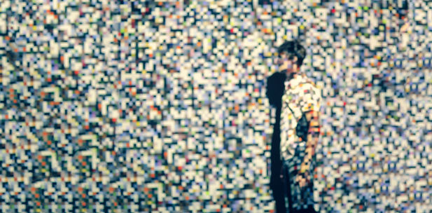 A young man stands against a multi-coloured heavily pixelated background.