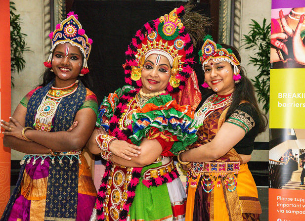 Three South Asian women smile with their arms crossed dressed in brightly coloured traditional celebratory dress.