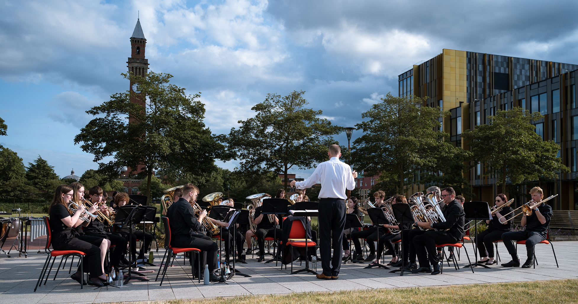 Members of the brass band performing outdoors with University of Birmingham buildings in the background