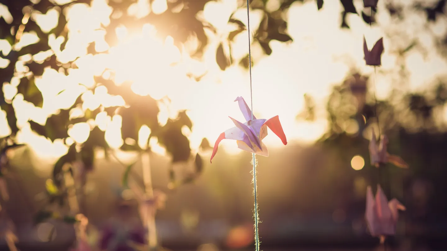 An origami bird hanging from string against a blurred background of leaves and sunlight.