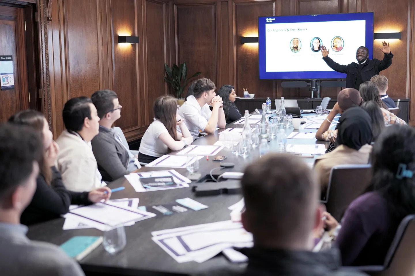 A man presenting to a long table of people in a wood-panelled boardroom with a screen behind him