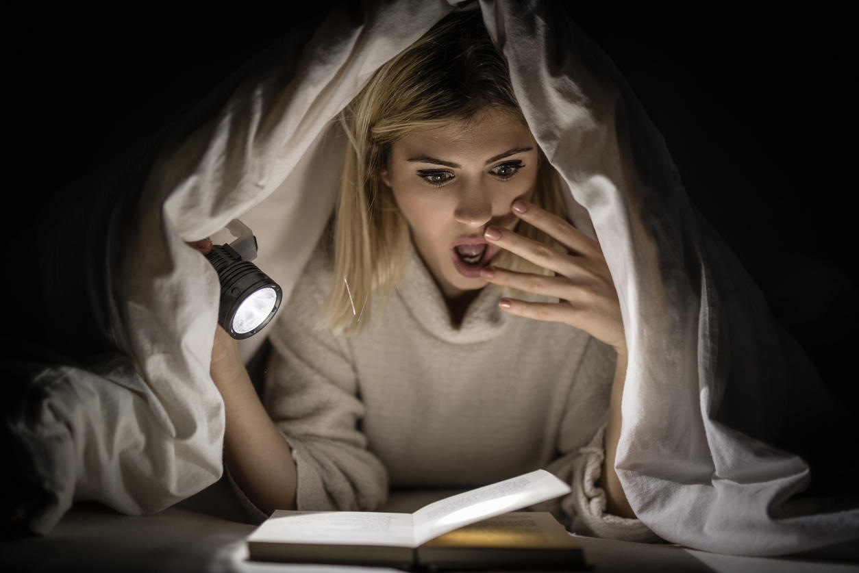 A women reading a scary book under the covers with a torch 