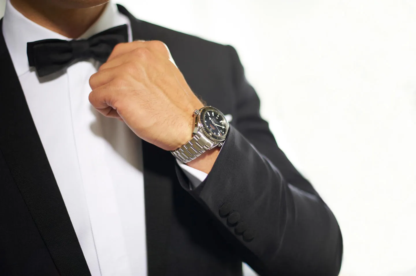 A man dressed in black dinner suit adjusting his bow tie, with a large chrome watch visible on his wrist