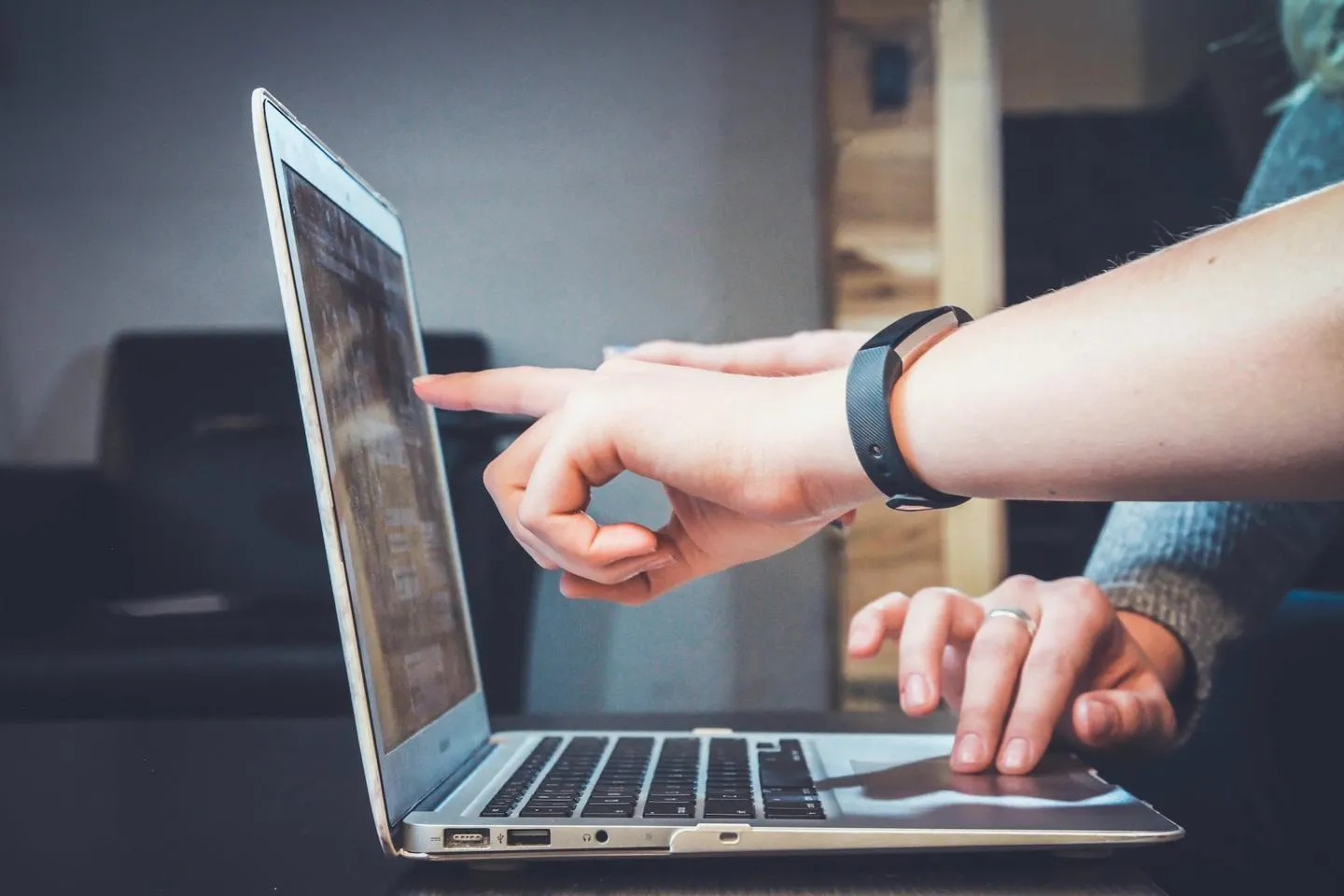 Two people pointing at and using touchpad of a laptop. One person is wearing fitness watch computer and other has grey long sleeved top