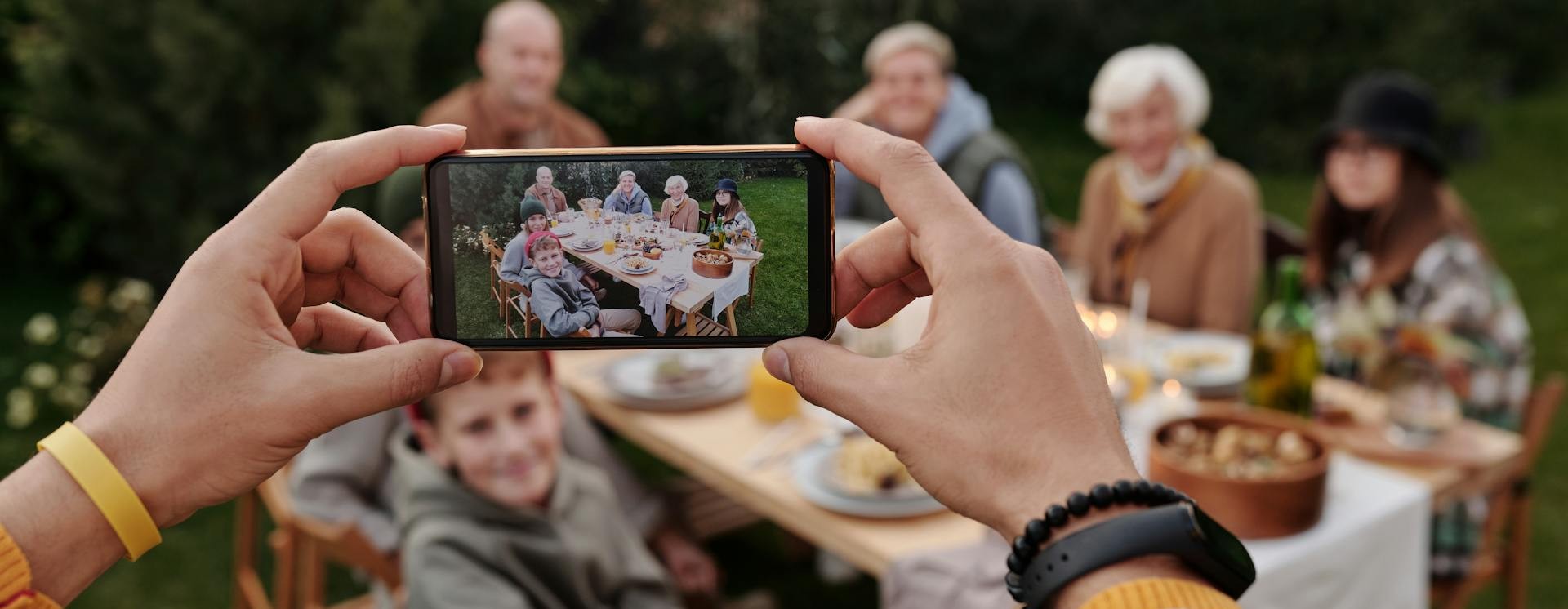 Hands holding up a smartphone that is taking a picture of a group sitting around a table in a garden