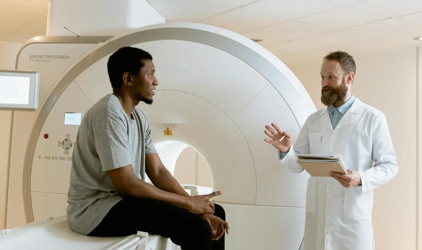 Clinician talking to young man sat on a bed with MRI scanner in background