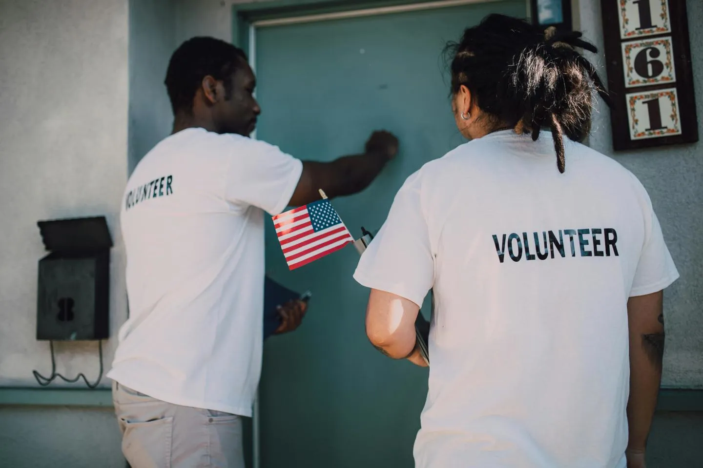 Two canvassers with volunteer t-shirts knocking on a door, one holding the American flag.