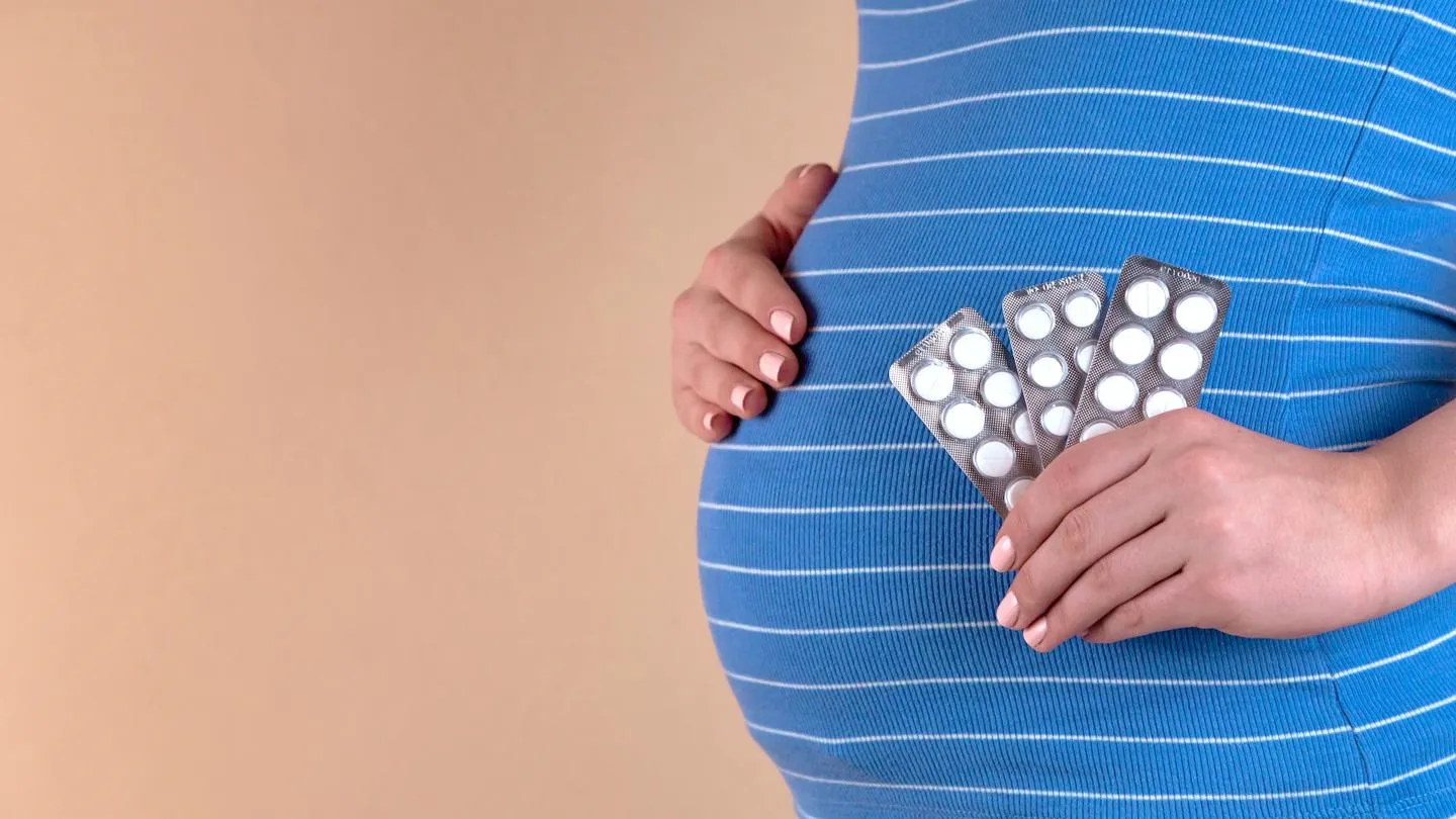  A close-up view of the belly of a pregnant woman in a blue T-shirt, which holds pills in her hands.