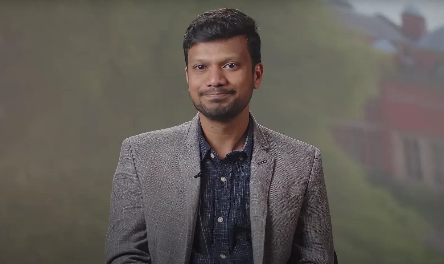 Dr Rohit Chikkaraddy in a blue shirt and grey jacket in front of an abstract background.