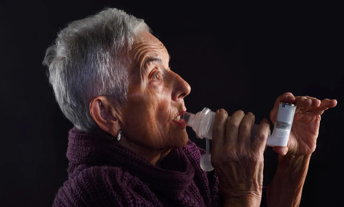 A woman with short grey hair and purple jumper using an asthma inhaler and spacer