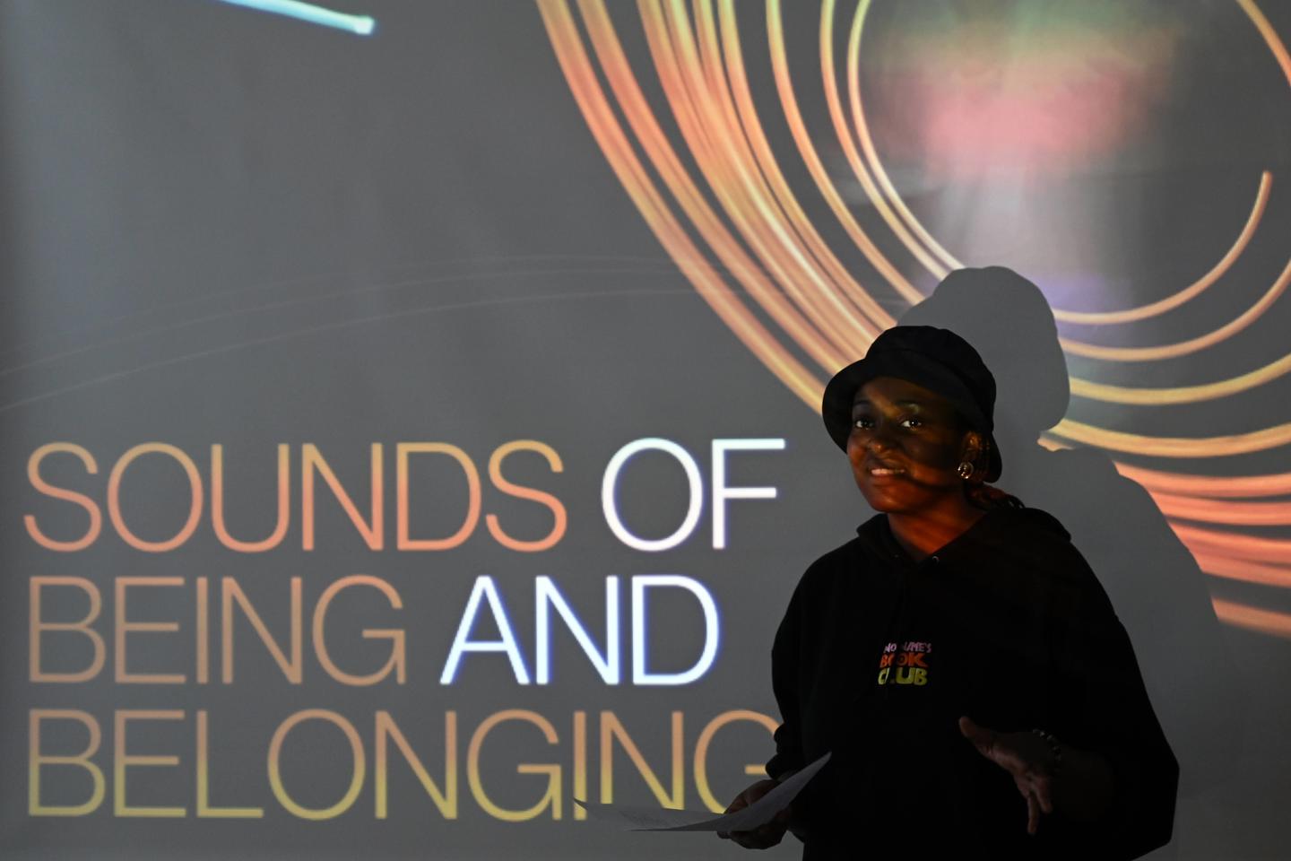 Artist Jaz Morrison talks in front of a wall with a projection reading 'Sounds of Being and Belonging'.