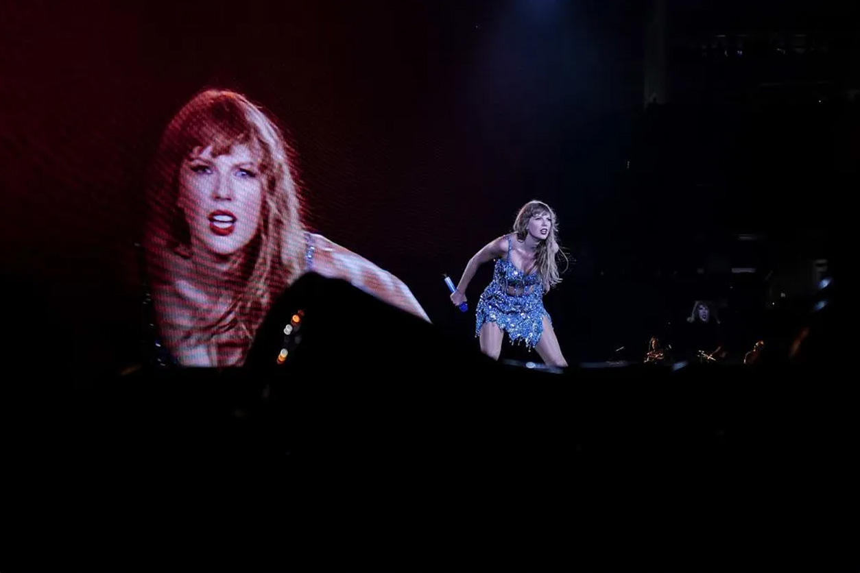 Taylor Swift performing at the Eras Tour, with her image projected onto a giant screen behind her