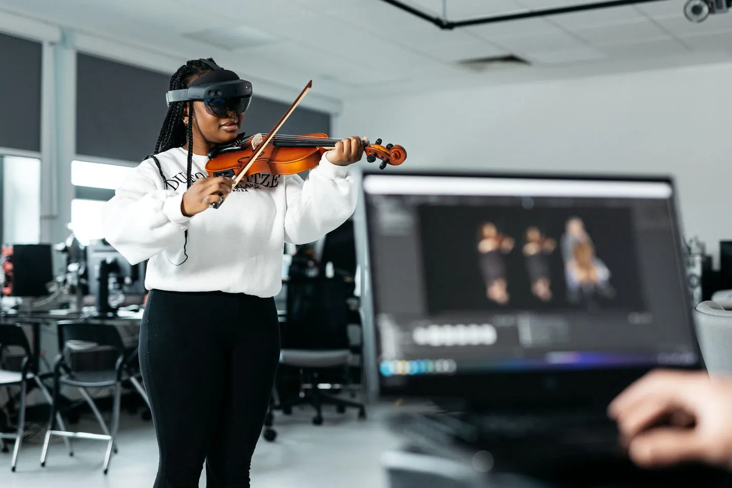 A woman wearing a VR headset plays the violin while someone looks at virtual musician avatars on a laptop in the foreground.