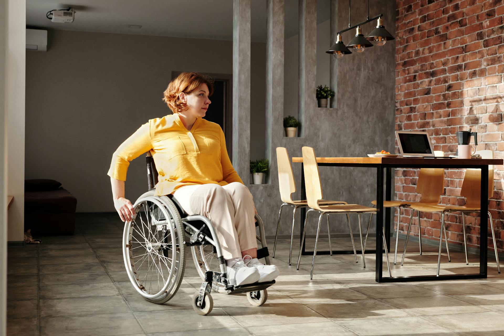 Woman wearing yellow shirt and neutral trousers in a wheelchair heading to laptop on dining table