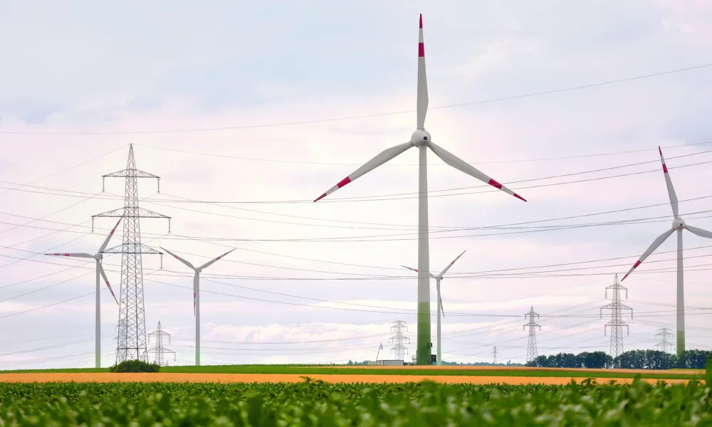 A cluster of wind turbines and pylons in a green and yellow field against a cloudy blue sky.