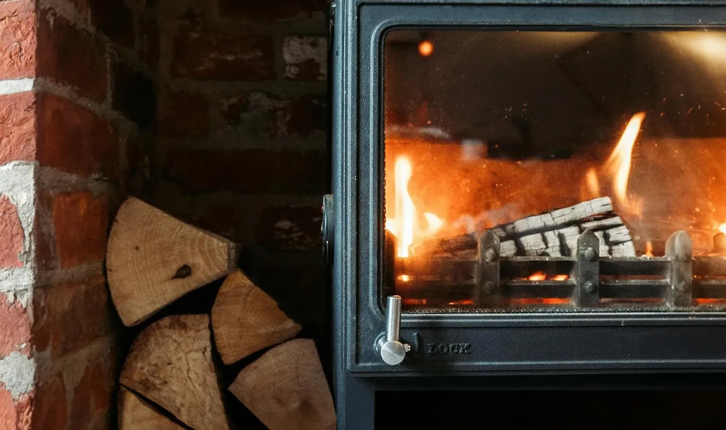 A woodburning stove in a brick alcove with logs aflame and a pile of fresh logs stacked beside it