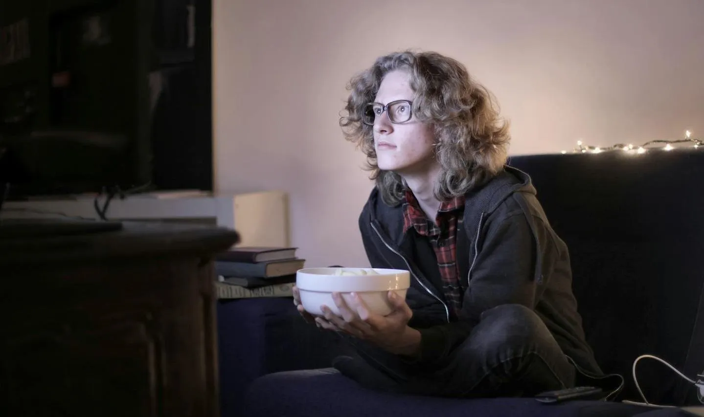 A young man wearing glasses watching television while holding a bowl of food