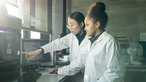 Two female students in labcoats pointing at computer screen with test results on