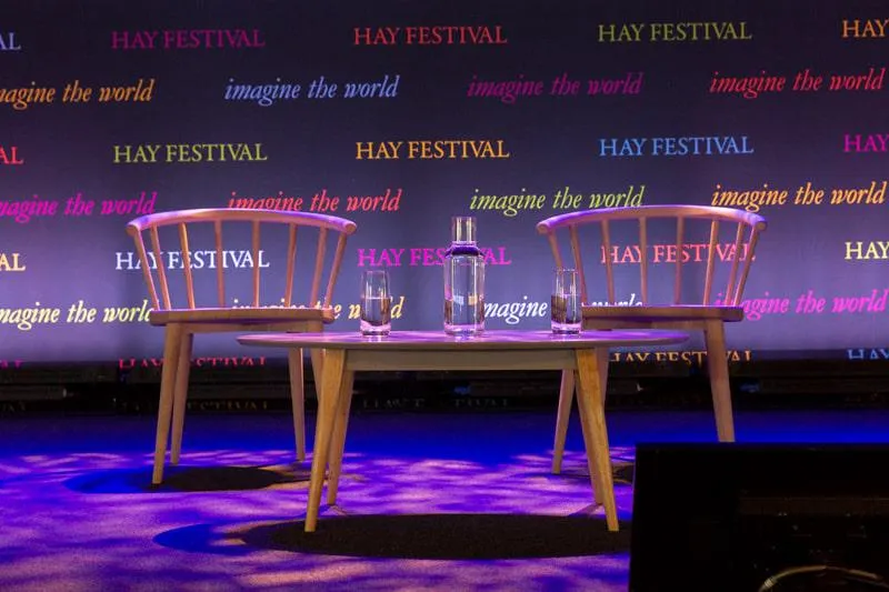 Two empty chairs and a small table with glasses of water on stage at the Hay Festival against a branded backdrop